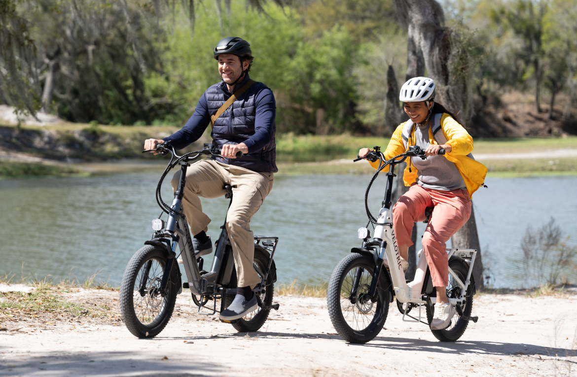 Two people riding Velotric Fold 1 eBikes along a lakeside path, smiling and enjoying a sunny day outdoors.