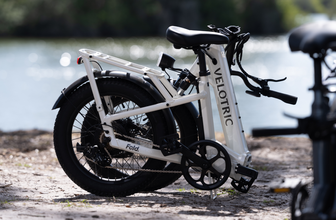 A Velotric Fold 1 eBike parked on a gravel path near a body of water, with the focus on its frame and wheels.