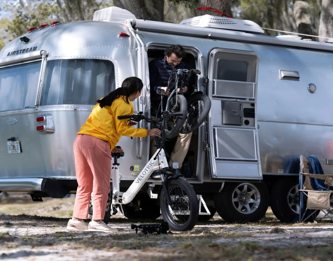 A man and a woman loading a Velotric Fold 1 eBike into an airstream trailer in a wooded area.