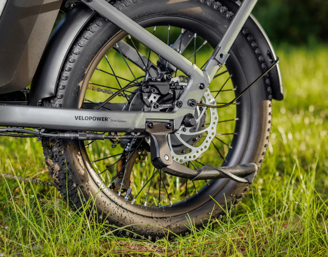 Close-up of a Velotric Fold 1 electric bike's rear wheel and motor, displaying its disc brake and thick tire on grassy terrain.