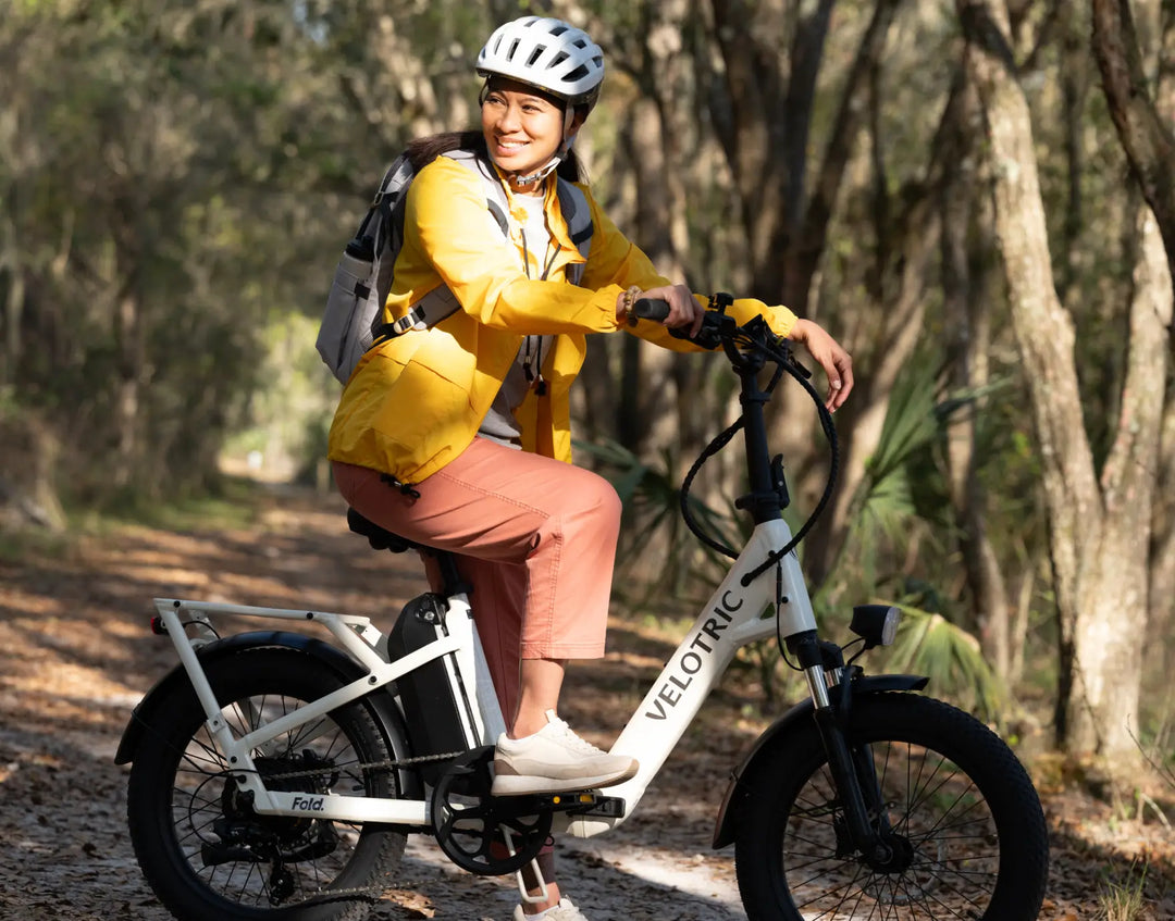 A woman in a yellow jacket and helmet smiling while standing next to a white Velotric Fold 1 eBike with a folding design on a wooded path.