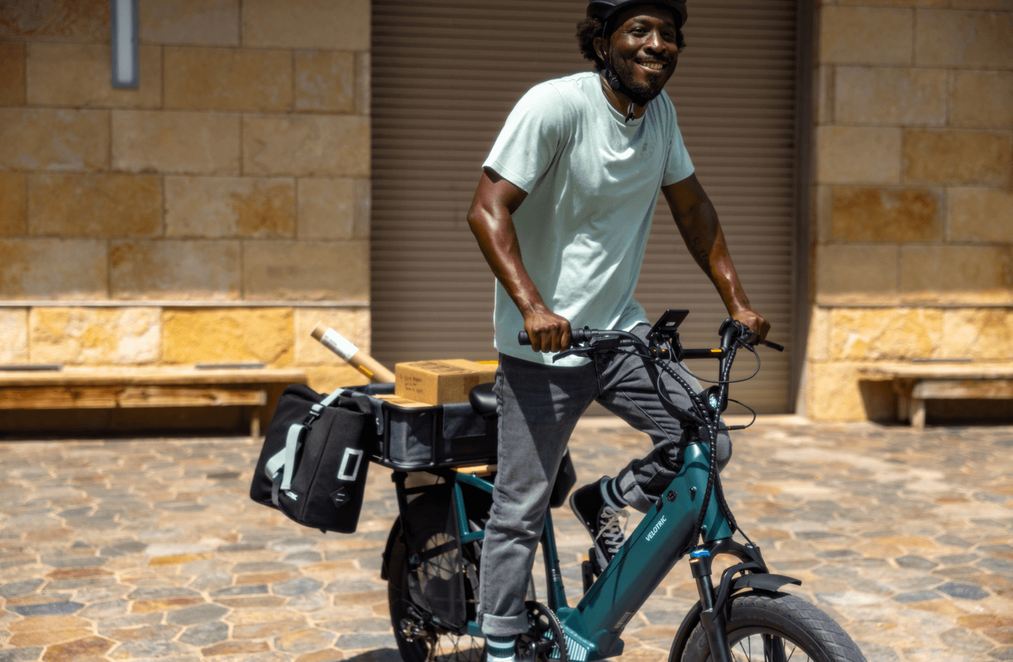 A delivery worker smiling while standing with a loaded Velotric - Go 1 - Forest utility eBike in front of a closed shop.