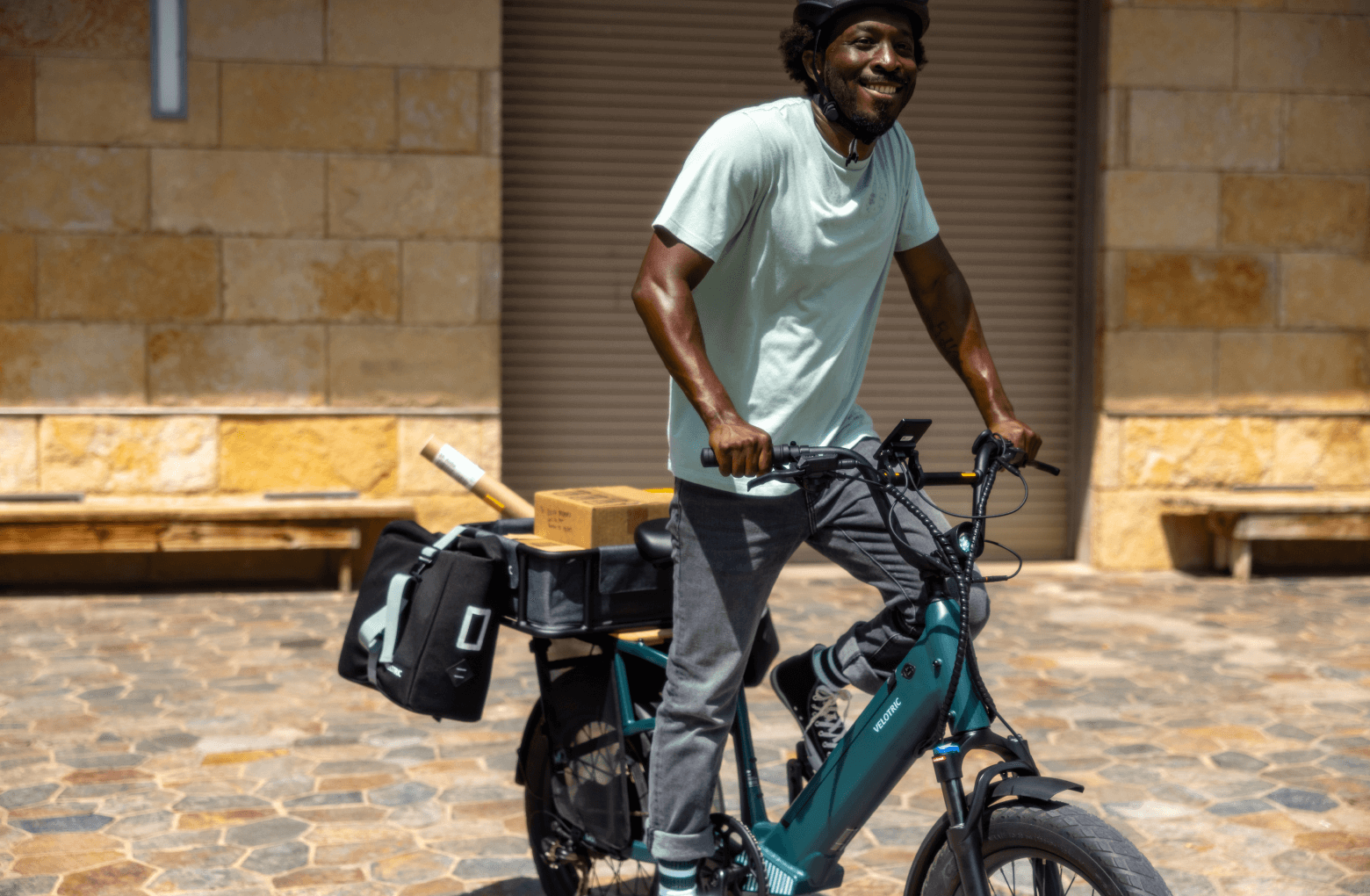 A delivery worker smiling while standing with a loaded Velotric - Go 1 - Forest utility eBike in front of a closed shop.