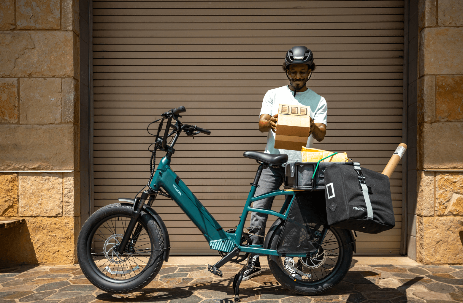 A delivery person checking a package beside a Velotric Go 1 - Forest foldable electric bike loaded with parcels, parked in front of a closed shutter.