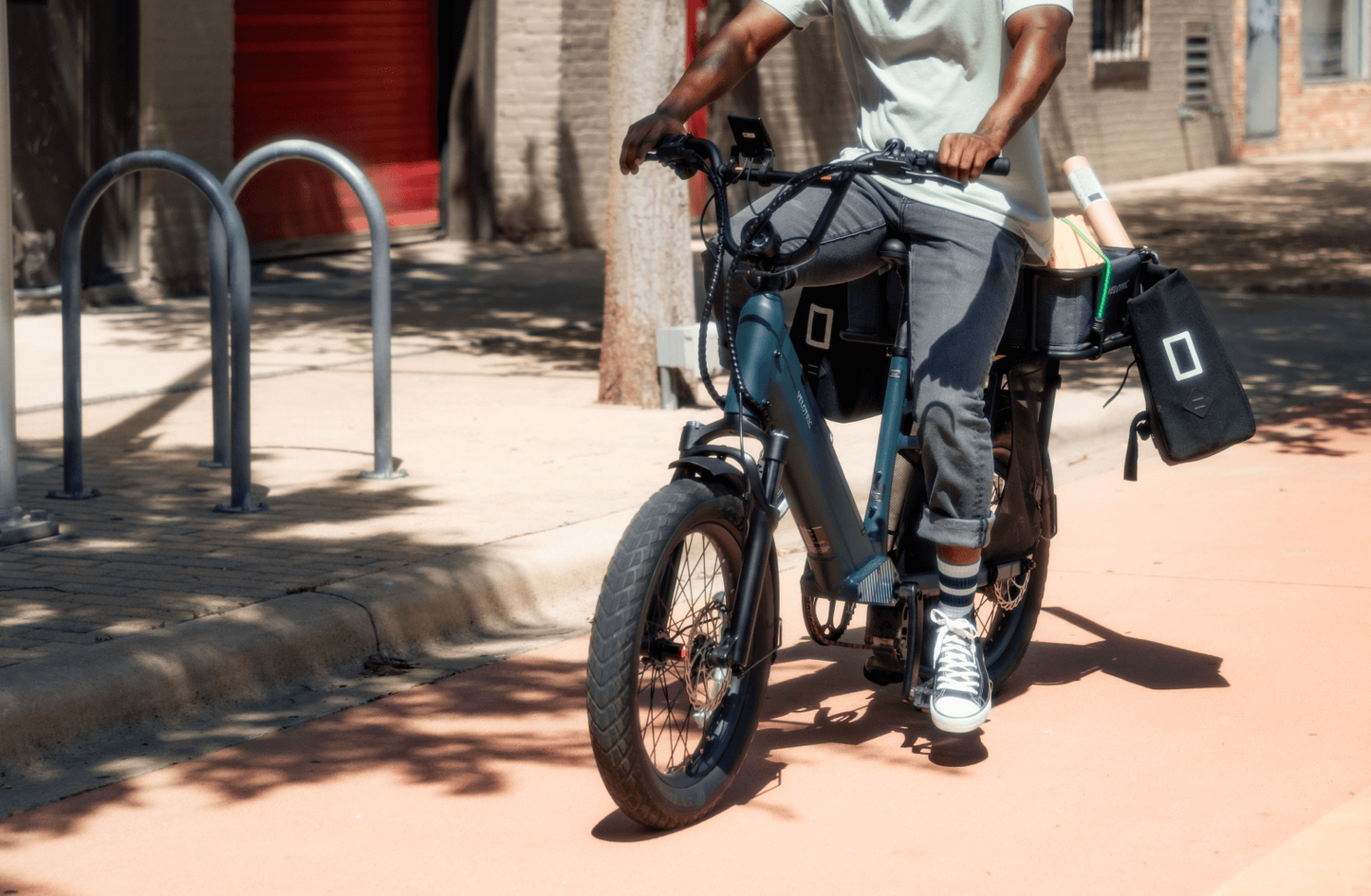 A person mounting a Velotric Go 1 - Forest electric bike parked on a sunny street, with a bag and books strapped to the bike.
