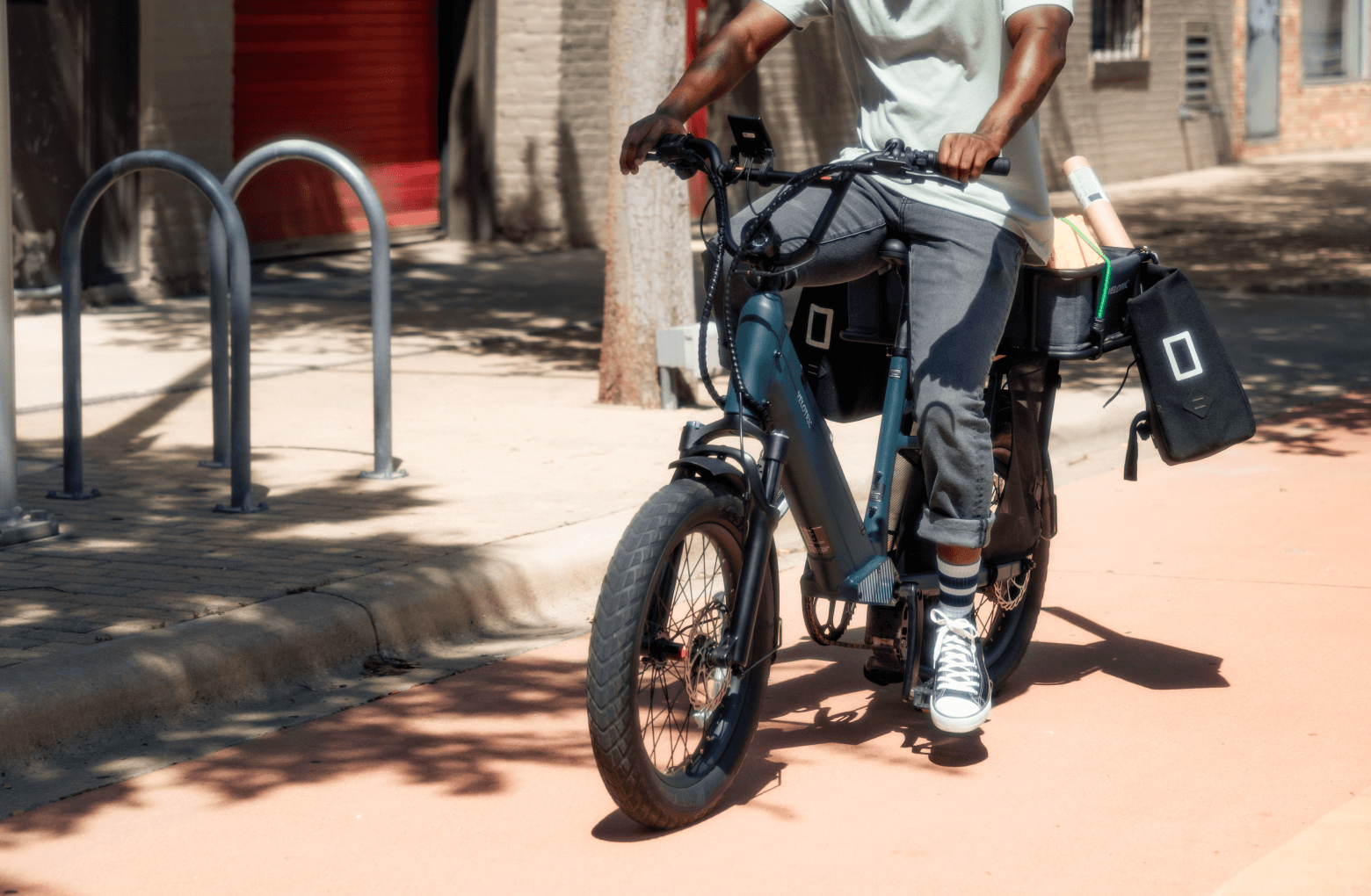 A person mounting a Velotric Go 1 - Forest electric bike parked on a sunny street, with a bag and books strapped to the bike.