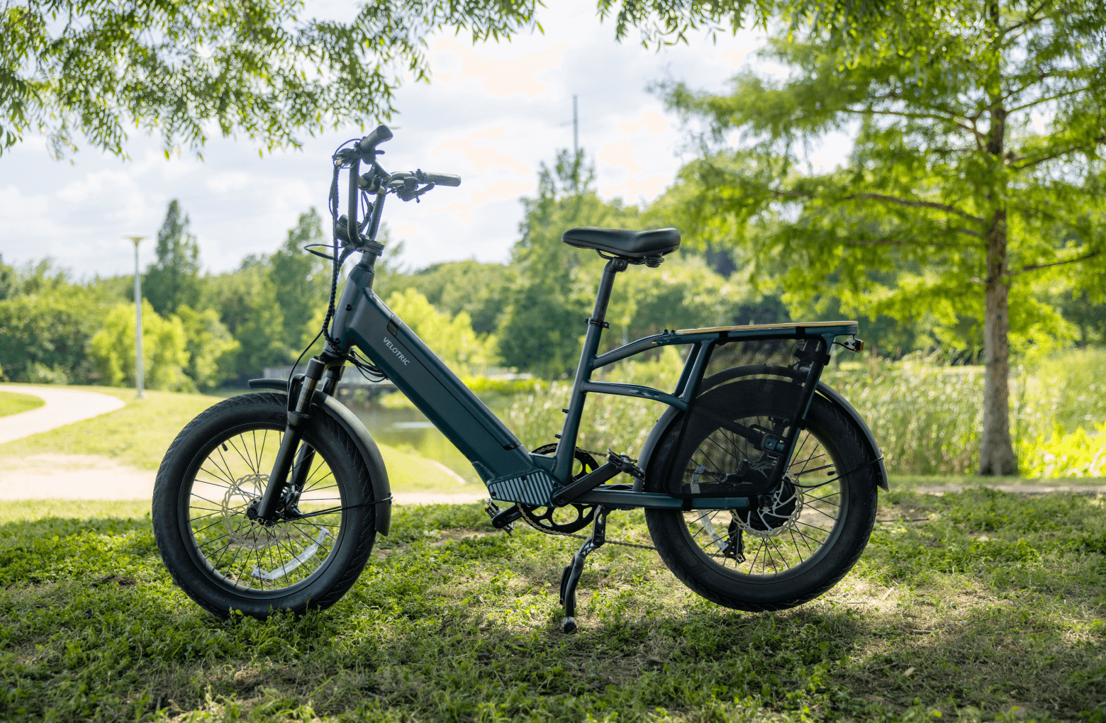 Electric bicycle, specifically a Velotric Go 1 - Forest, parked on grass with trees and a path in the background on a sunny day.