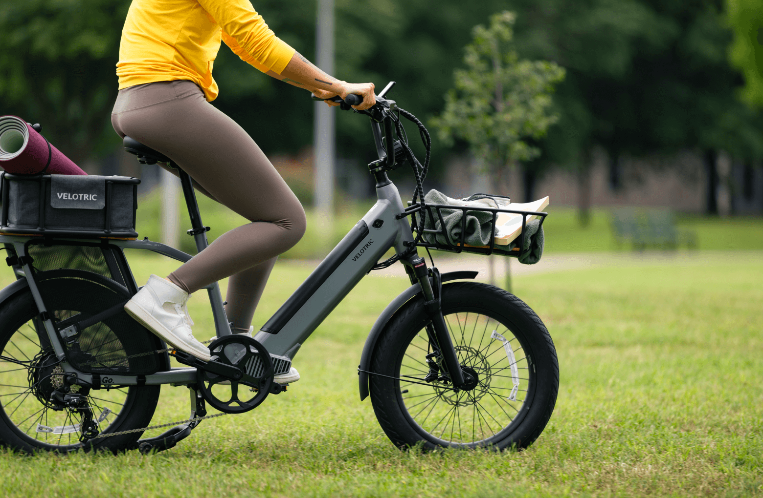Woman in a yellow shirt and brown leggings mounting a Velotric Go 1 electric bike with a rear carrier in a park setting.