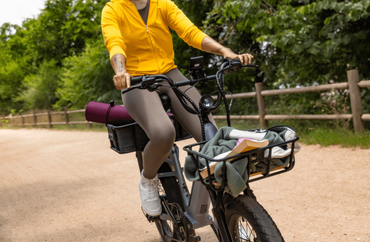 A person in a yellow jacket rides a Velotric Go 1 Forest electric bike on a dirt path, with yoga mat and towel attached.