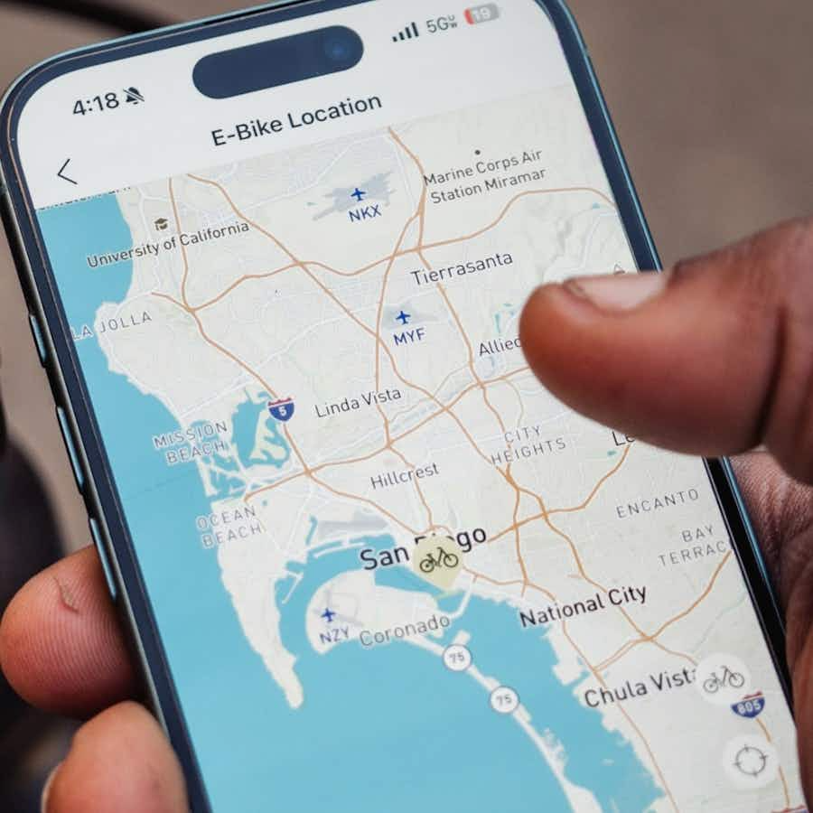A hand holds a smartphone showing a map of San Diego with various Aventon - Aventure 3 e-bike locations from Aventon marked on the screen.