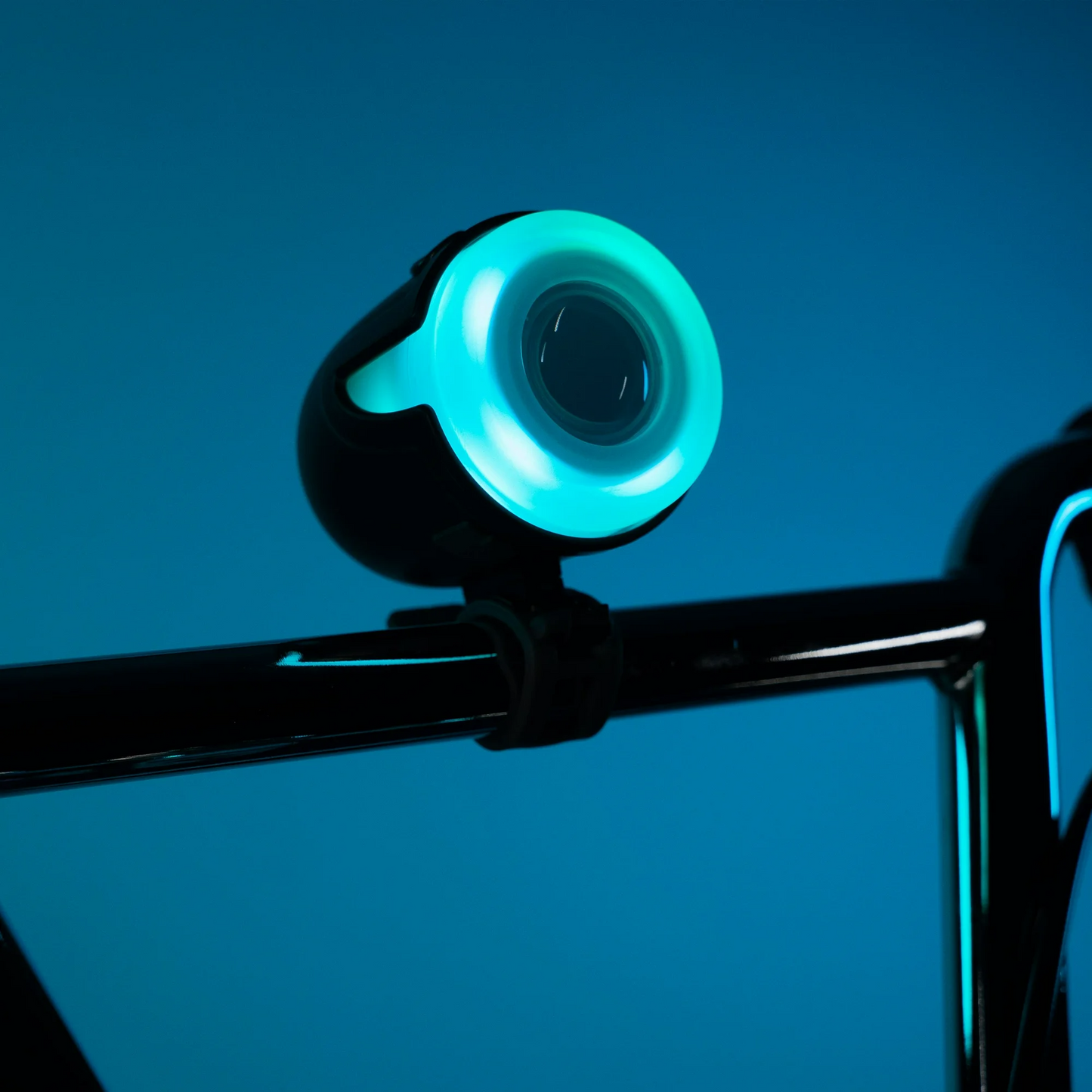Close-up of a weather-resistant Brightz Halo Brightz bicycle headlight emitting a blue-green glow, mounted next to an LED light-up bell on black handlebars with a blue background.