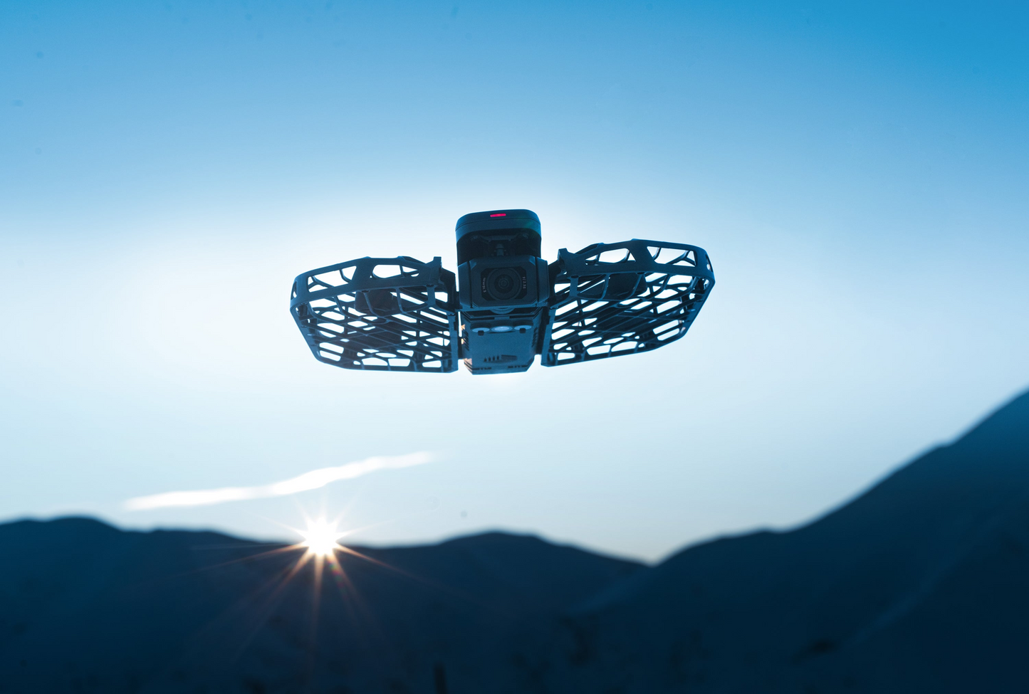 The HOVERAir X1 Pro Max by HOVERAir, a self-flying camera with AI tracking, elegantly flies against the backdrop of a beautiful mountain sunset, its propeller guards silhouetted in the sky.