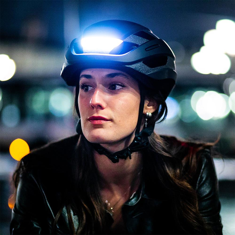 Woman wearing a Lumos Ultra MIPS Helmet with integrated LED lighting at night.