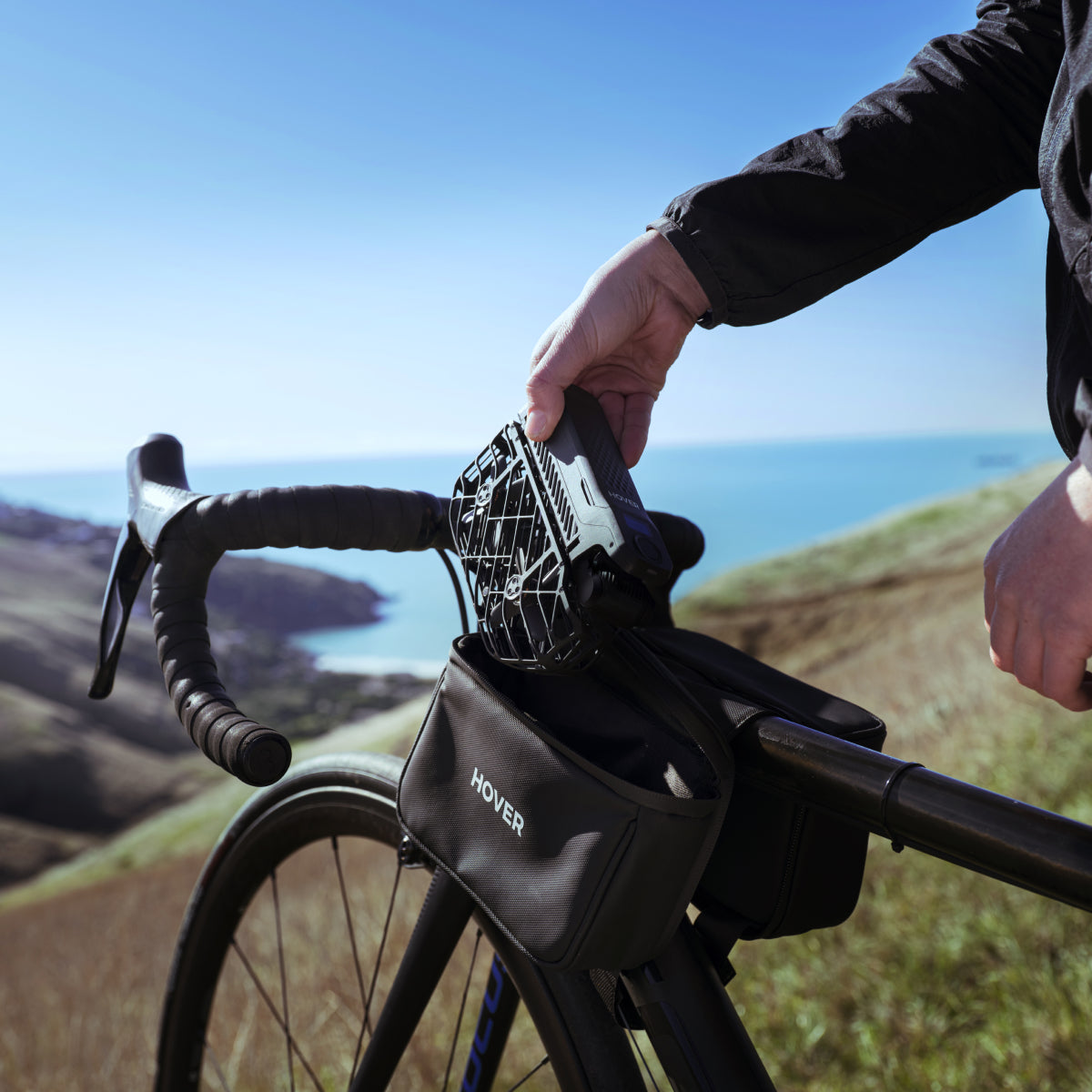A person places a black speaker into a HOVERAir Carry Bag inside a handlebar bag on their bicycle, overlooking a scenic coastal landscape.
