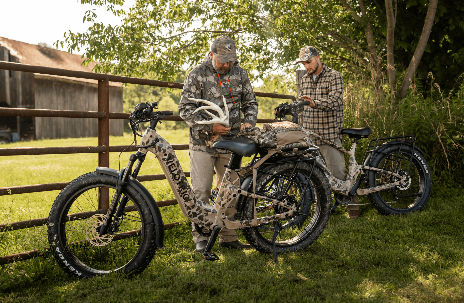 Two men in camouflage stand beside two Velotric - Nomad 2X fat tire eBikes from Velotric near a metal fence, examining outdoor gear on the grass.