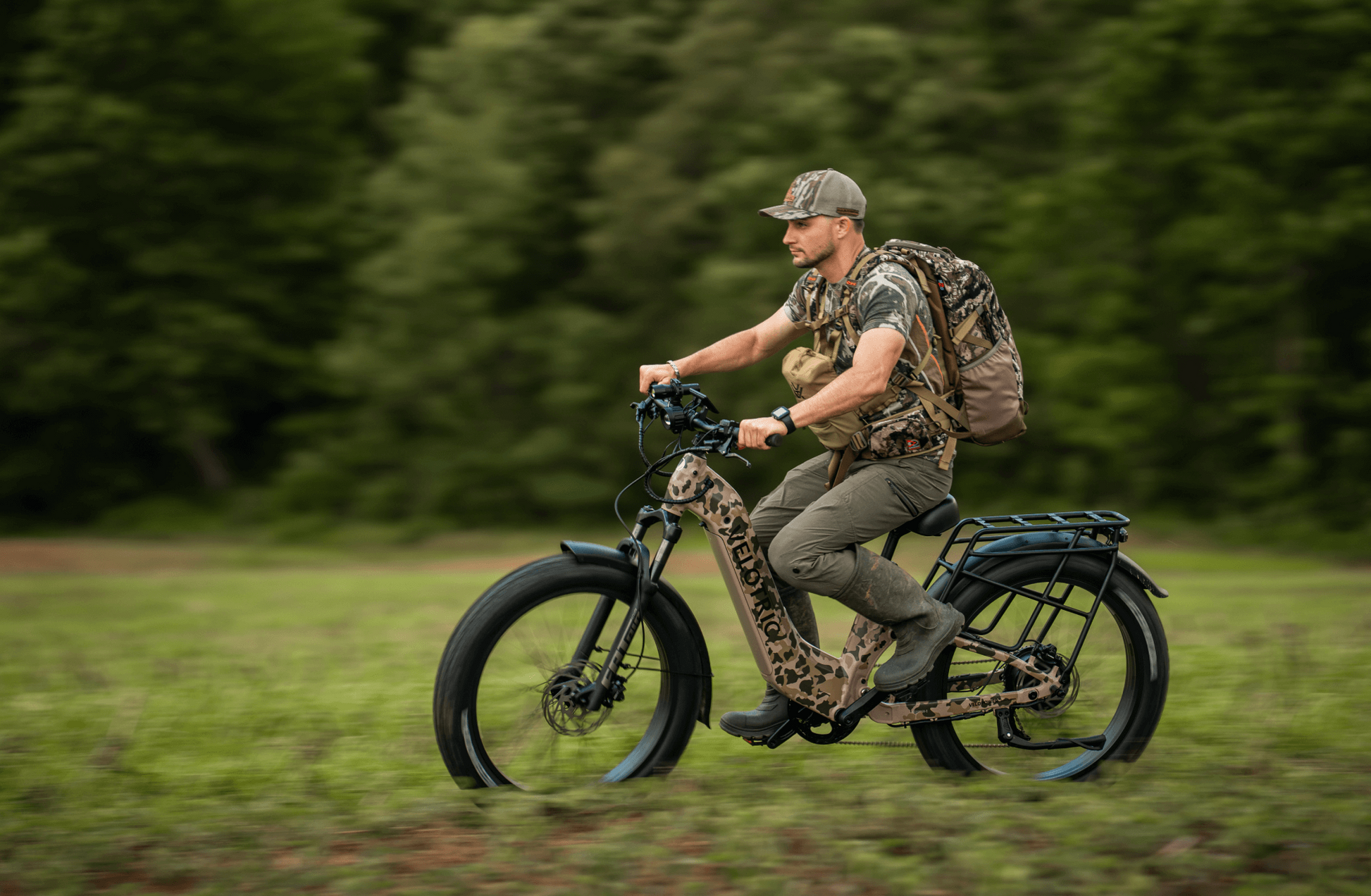 A man in camouflage clothing and a backpack rides the Velotric - Nomad 2X full suspension eBike by Velotric, featuring a camouflage pattern, across a grassy field with trees in the background.