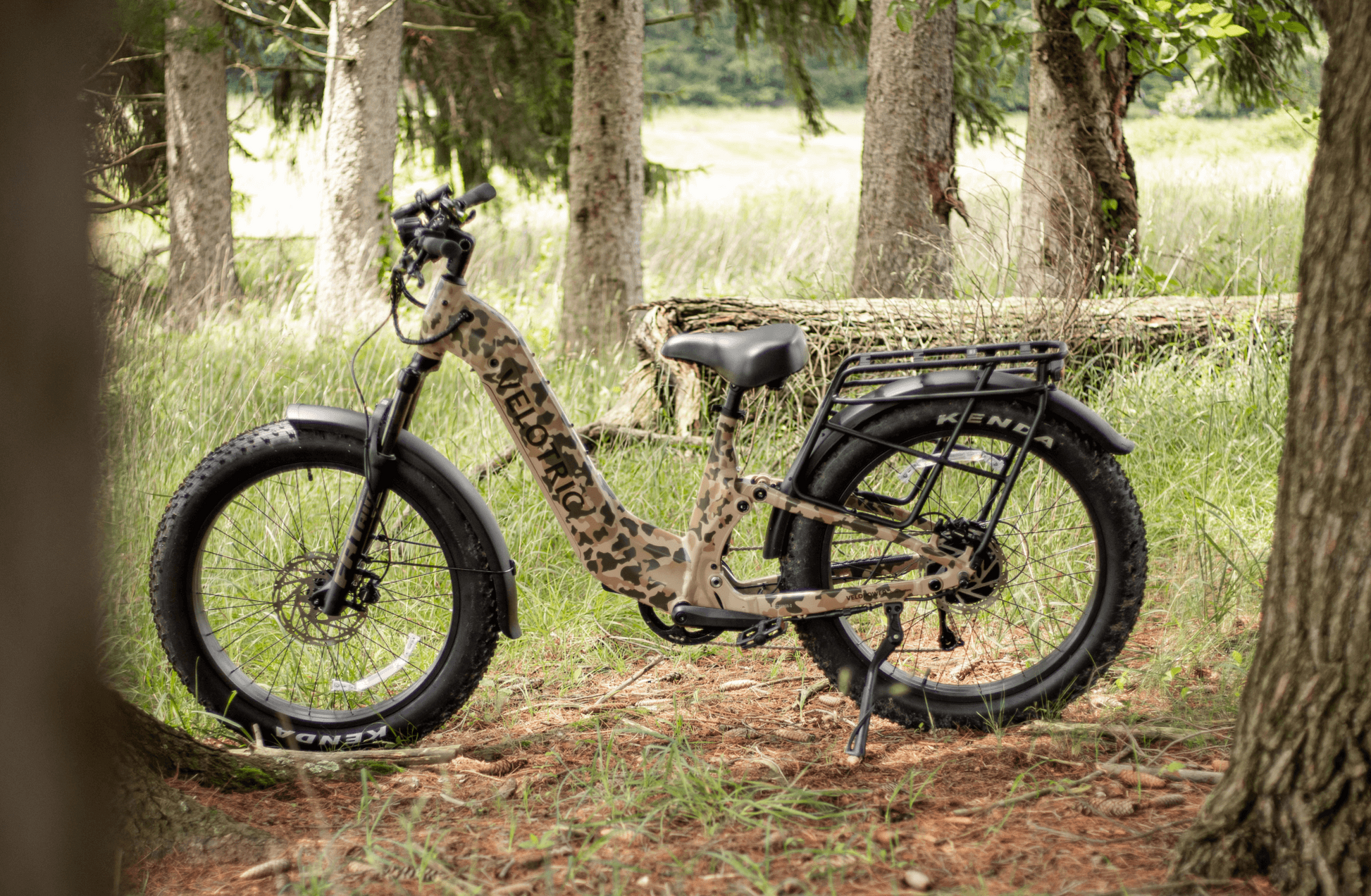 The Velotric Nomad 2X, a camouflage-patterned fat tire eBike with full suspension, is parked on grassy ground near trees and a fallen log.