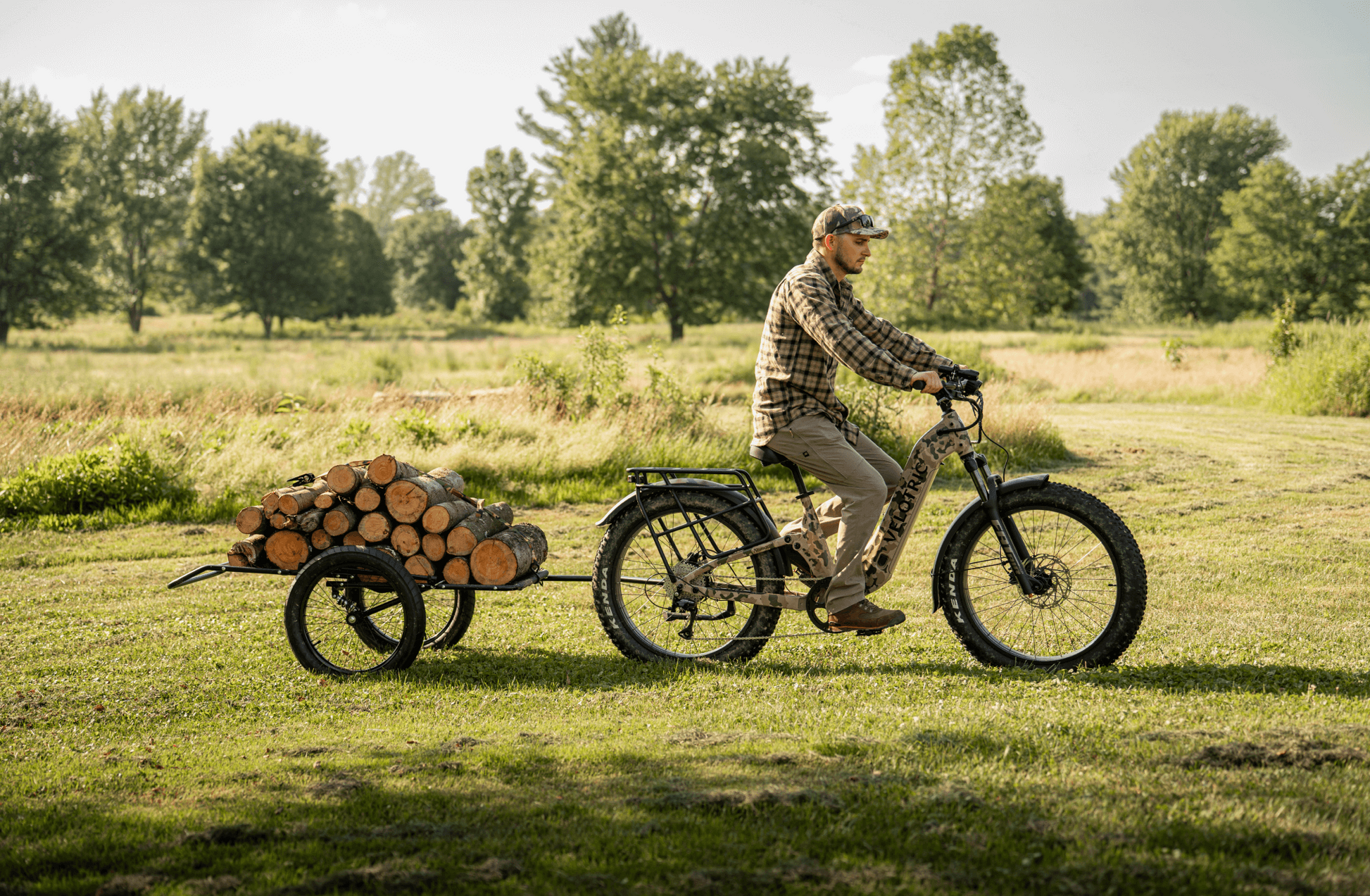 A man rides the Velotric Nomad 2X fat tire eBike by Velotric, towing a small trailer full of cut logs across a grassy field with trees in the background.