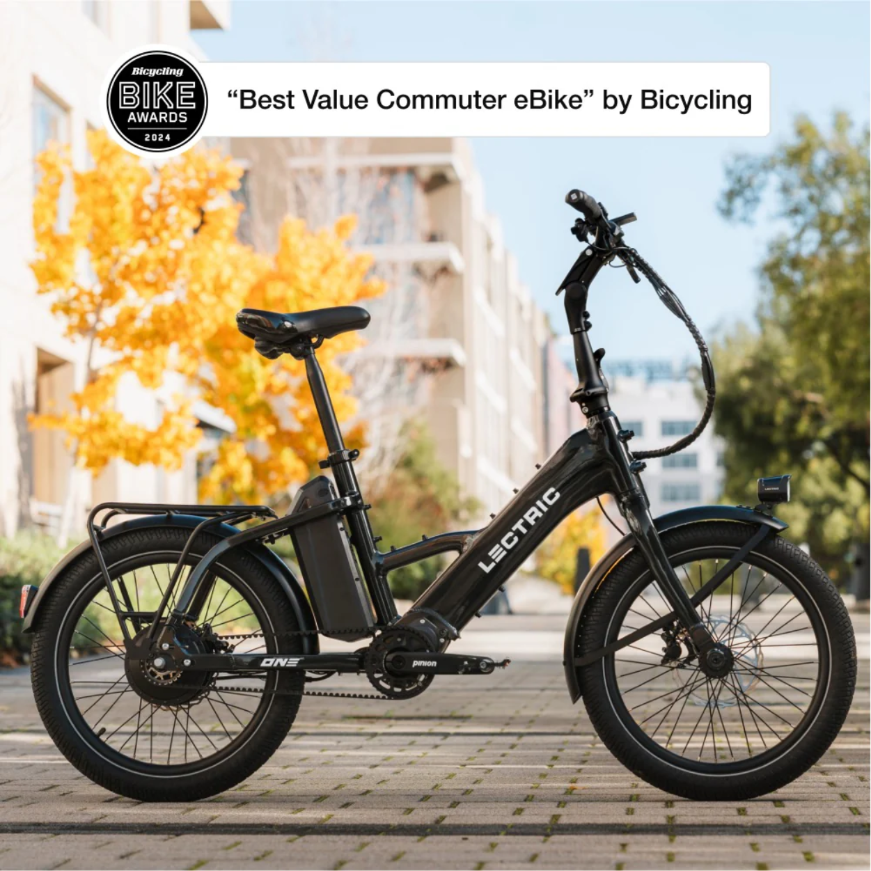 A black Lectric - ONE eBike stands on a paved path with buildings and a yellow-leaved tree behind it. A badge shows it won the 2024 “Best Value Commuter eBike” award.
