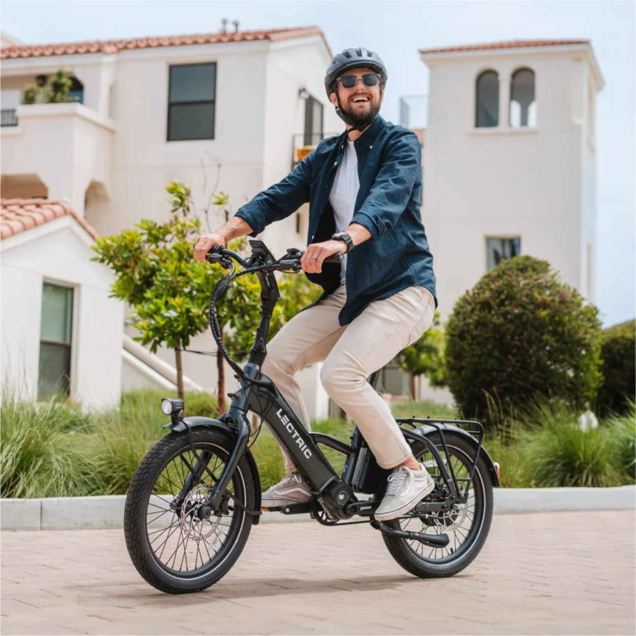 A man in a helmet, sunglasses, and casual clothes rides a black Lectric - ONE electric bicycle by Lectric on a paved path in front of white residential buildings.