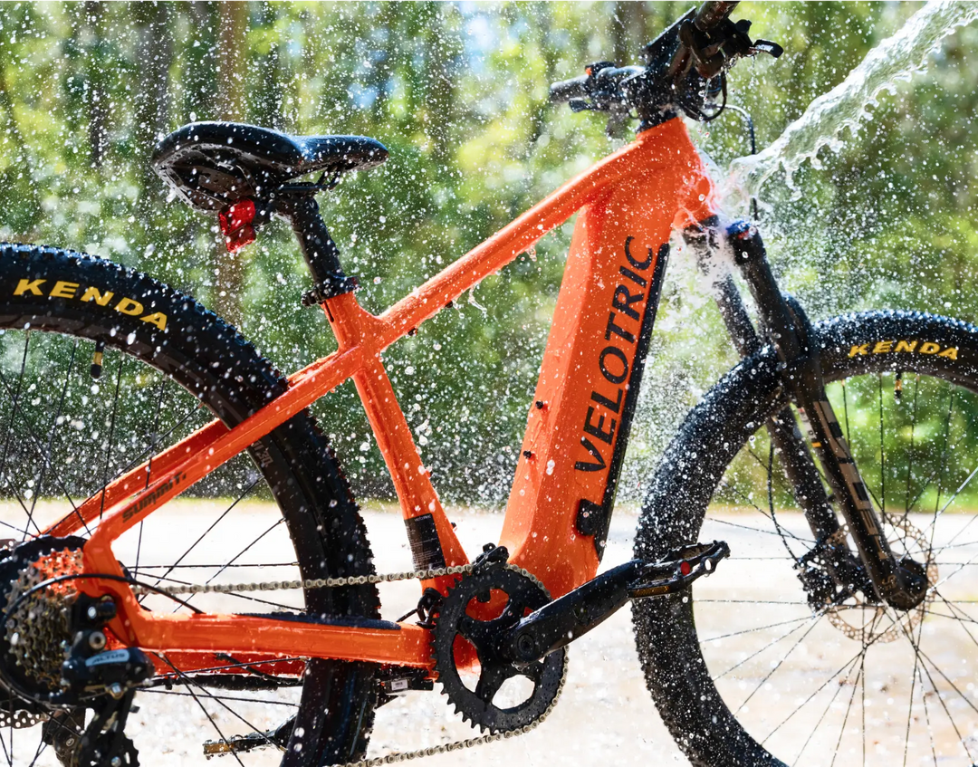 Orange Velotric Summit 1 eBike being washed outdoors with a spray of water droplets visible in the air, set against a blurred natural background.