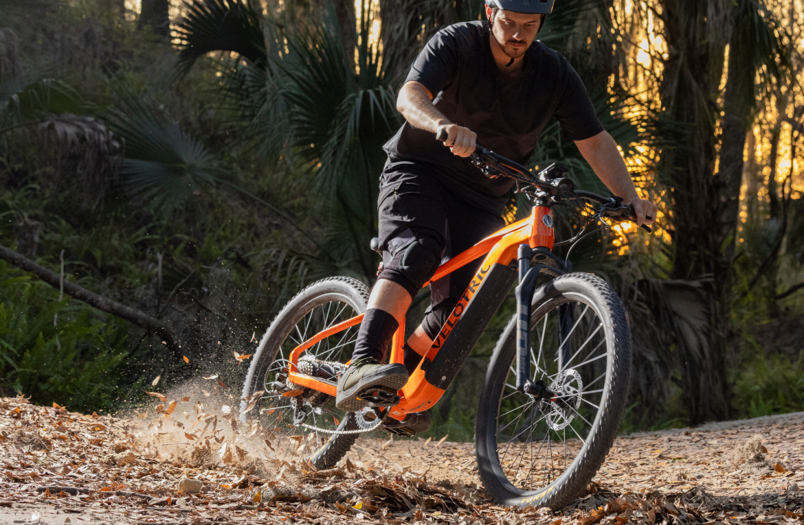 A cyclist wearing black shorts and a black cap rides a Velotric Summit 1 eBike on a dirt trail, kicking up leaves and dirt at sunset.
