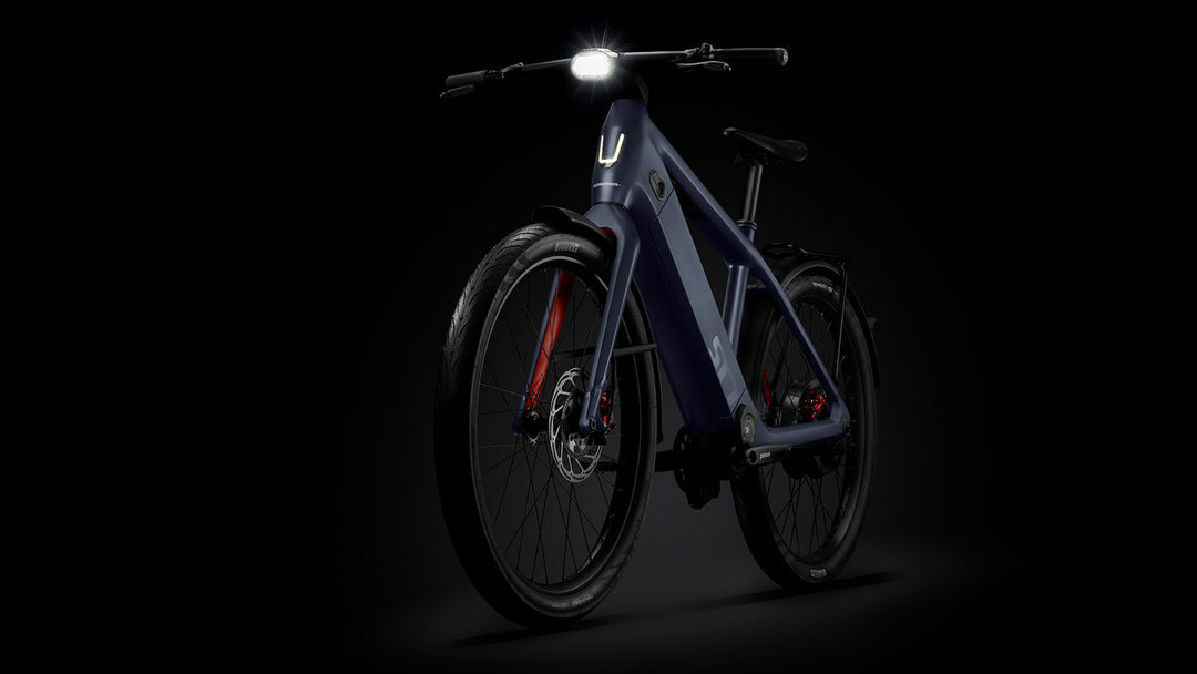 Electric bicycle with metallic blue frame illuminated by its front light, displayed against a dark background, Stromer - ST7 Alinghi Red Bull Racing Edition.