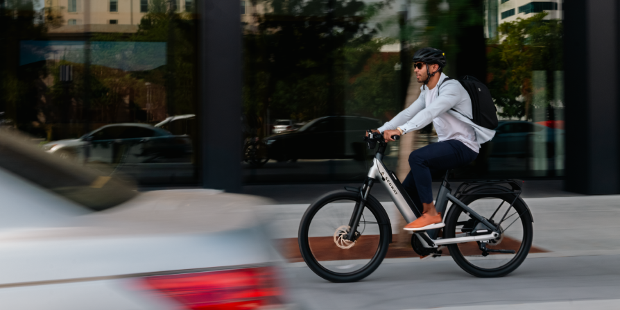 A man wearing sunglasses and a backpack rides the Segway - Myon by Segway, a popular commuter eBike with an 80-mile range, on a city street while blurred cars pass in the foreground.