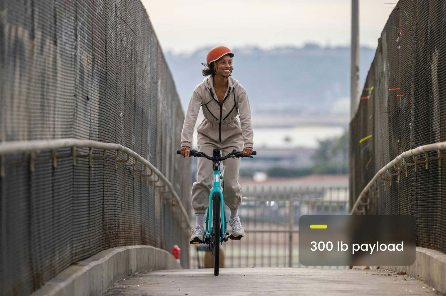 A person in an orange helmet rides a teal Aventon Soltera 2.5 bike with an aerodynamic frame across a fenced pedestrian bridge; text overlay states, "300 lb payload.