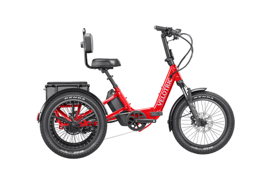 The Velotric Triker is a red electric tricycle featuring two rear wheels, a backrest seat, and a battery behind the seat. The brand name "Velotric" appears on the frame, offering powerful urban eBike performance.