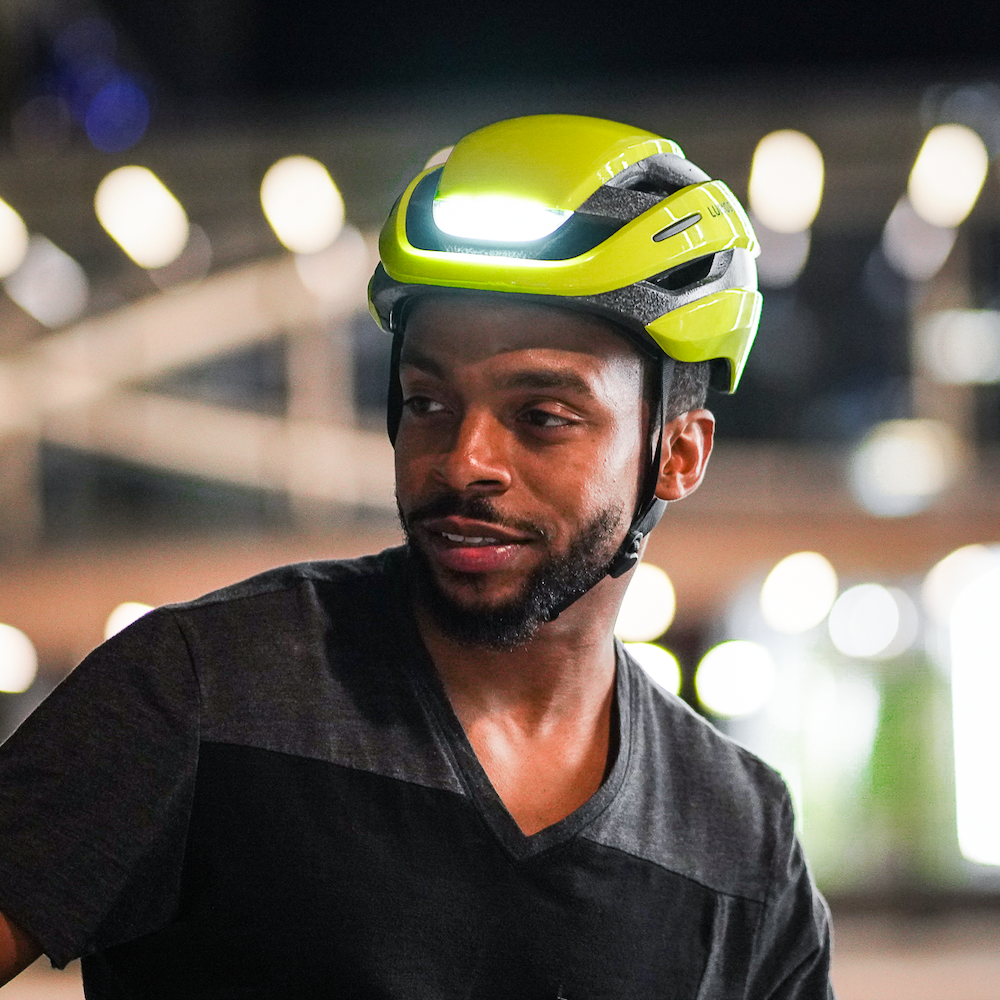 A man wearing a Lumos Ultra MIPS Helmet looks to the side with a thoughtful expression, possibly taking a break during a night cycling session.