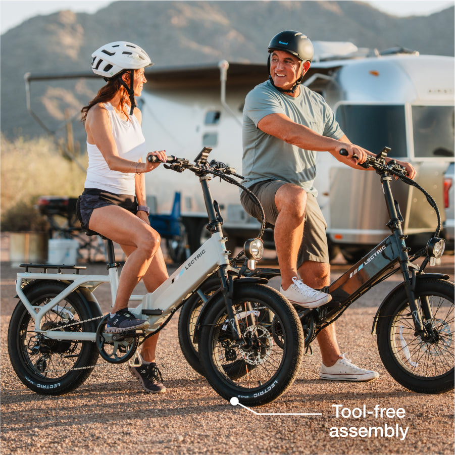 Two people wearing helmets sit on Lectric XP4 folding eBikes in a sunny outdoor setting with mountains and an RV behind them. Text on image reads: "Tool-free assembly.