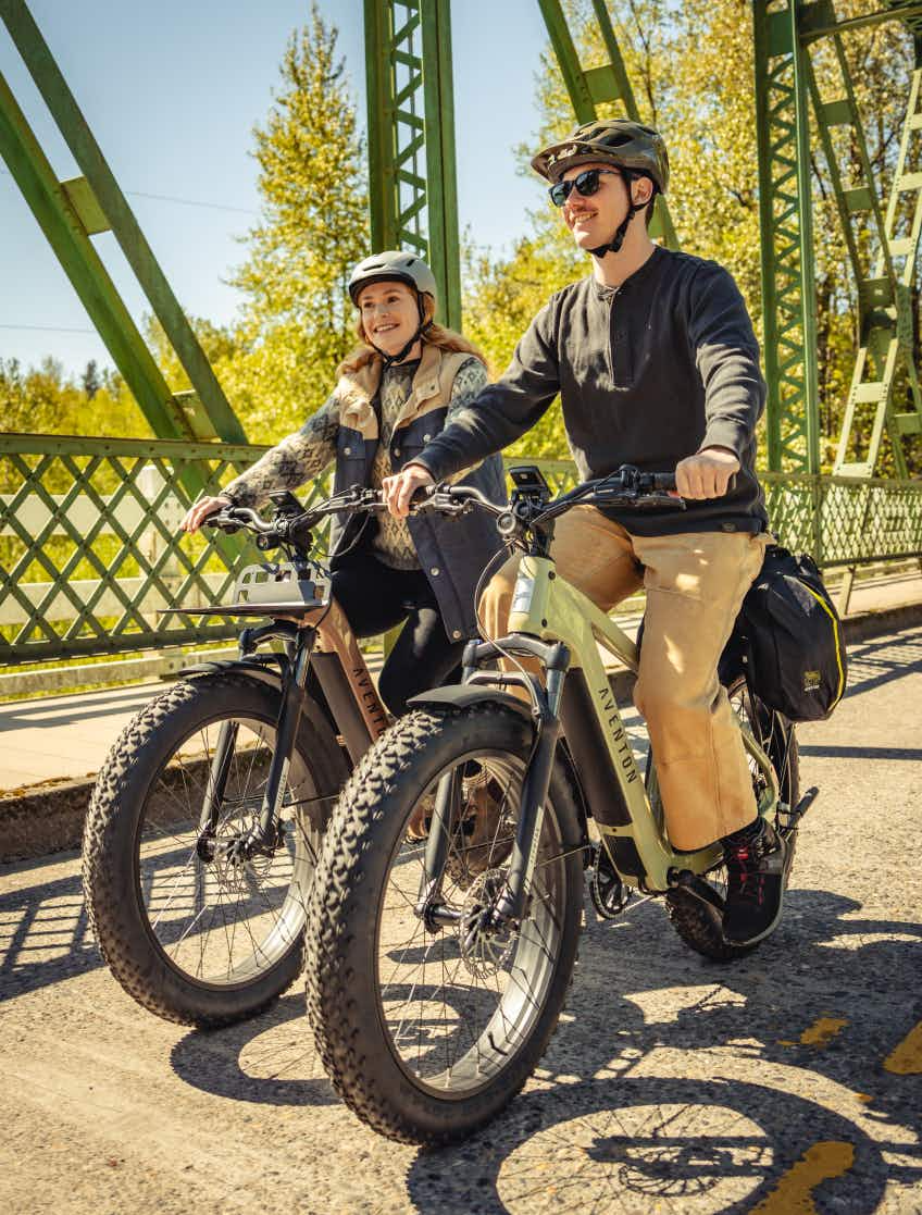 Two people wearing helmets ride Aventon Aventure 3 eBikes from Aventon side by side on a sunny day across a green metal bridge.