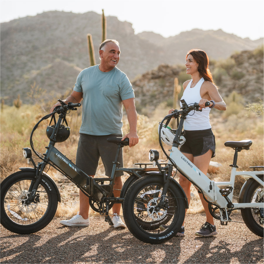 Two people with Lectric XP4 folding eBikes smile at each other on a desert trail surrounded by cacti and mountains—ready to ride with up to an 85-mile range from Lectric.