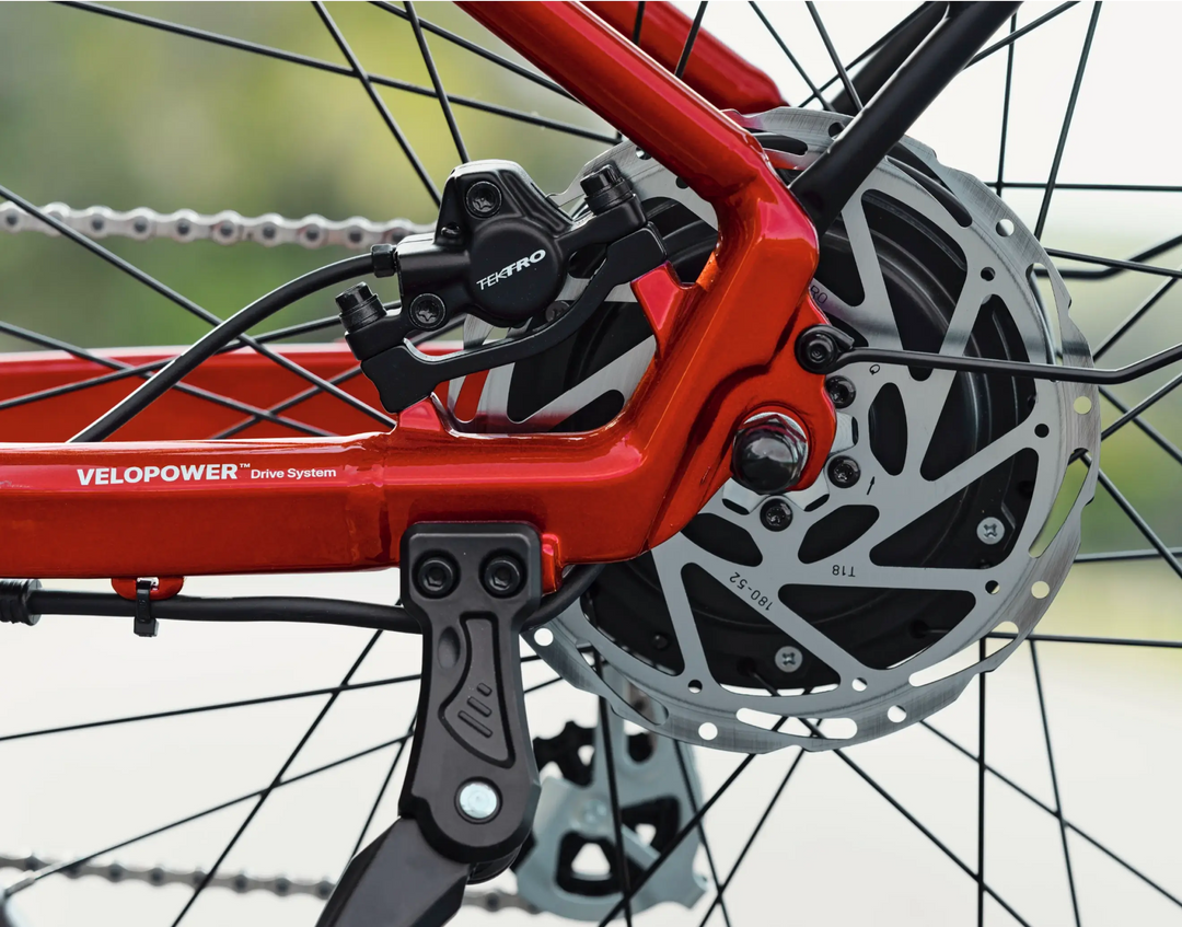 Close-up of the rear wheel of a red Velotric - Discover 1 electric bike showing the disc brake system, with visible branding "VeloPower" and "Tektro", available at Tampa Bay eBikes.