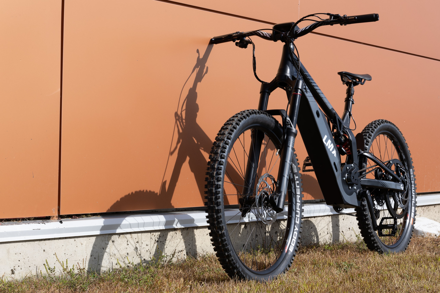 A black LMX - 64 electric bike by LMX with thick tires is parked on grass against an orange wall, casting a clear shadow—one of Tampa Bay eBikes' standout models.