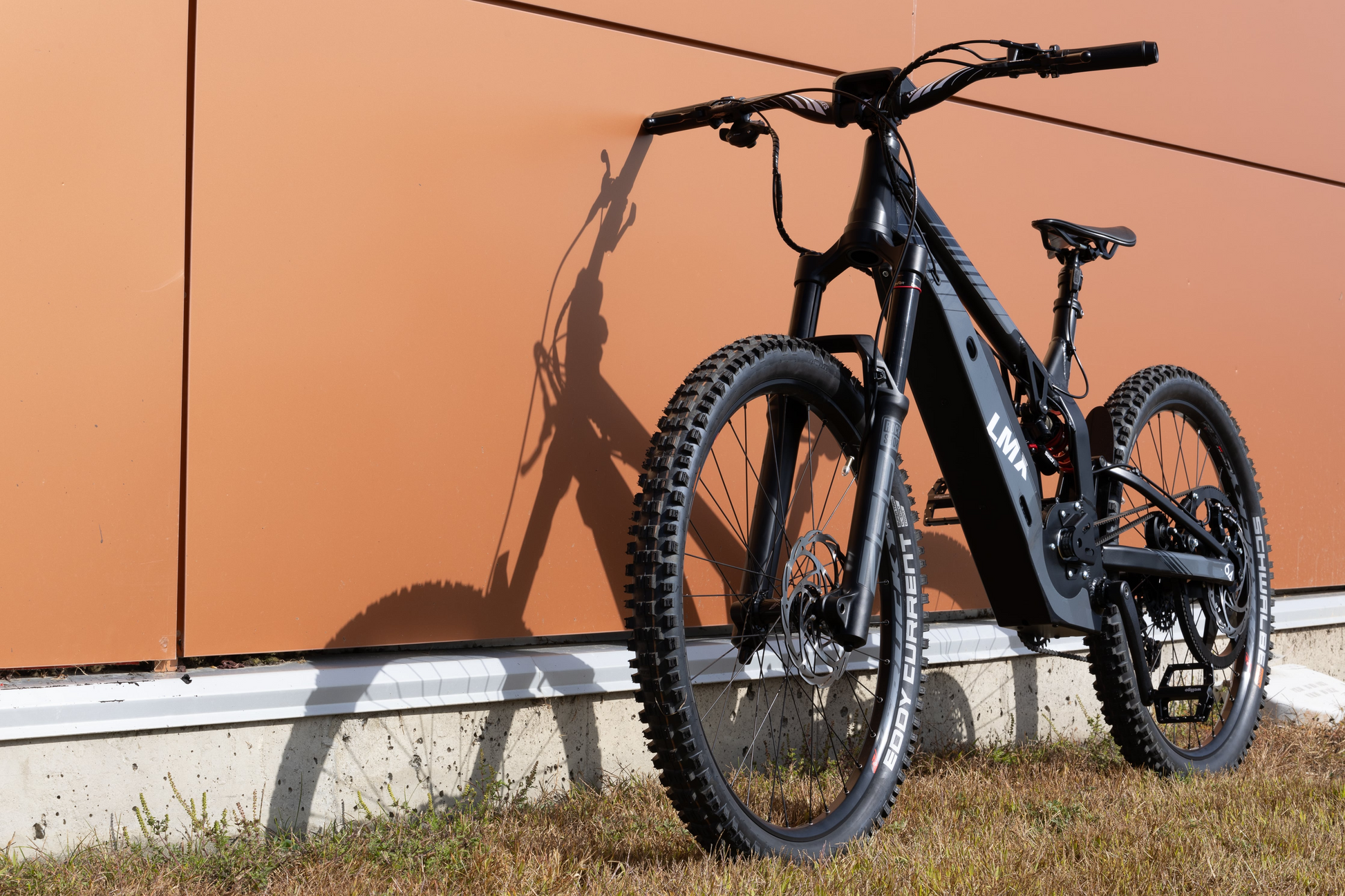 A black LMX - 64 electric bike by LMX with thick tires is parked on grass against an orange wall, casting a clear shadow—one of Tampa Bay eBikes' standout models.