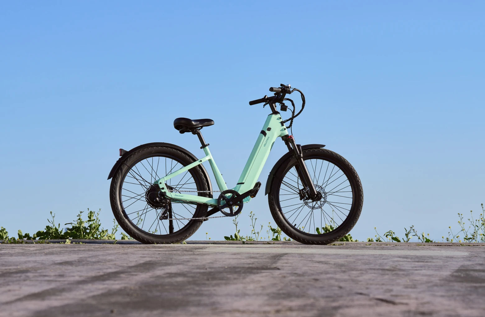 A light green Velotric - Discover 1 Plus commuter e-bike stands on a paved surface, featuring the powerful Velopower H50 drive system, with a clear blue sky in the background.
