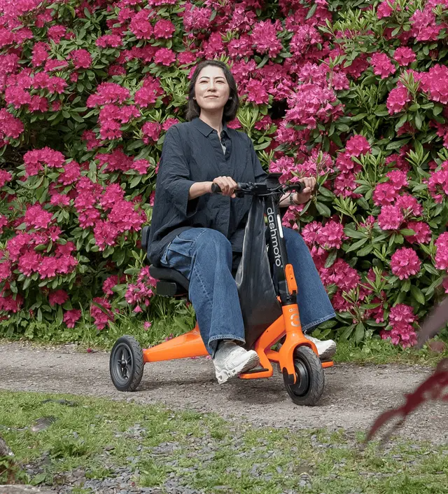 A person rides a Dashmoto Dash 3 orange folding mobility scooter by a path lined with a large bush of bright pink flowers.