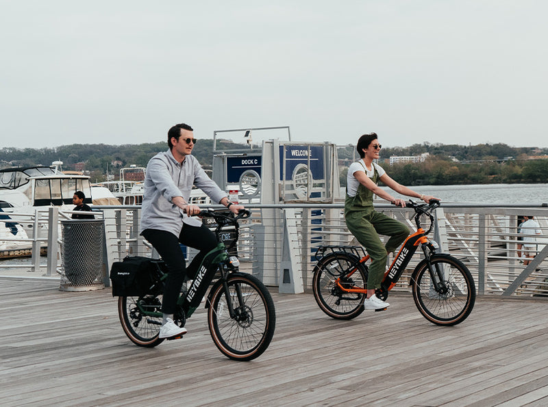 Two people riding HeyBike - Cityrun ebikes with hydraulic front suspension on a wooden pier, with boats and water in the background.