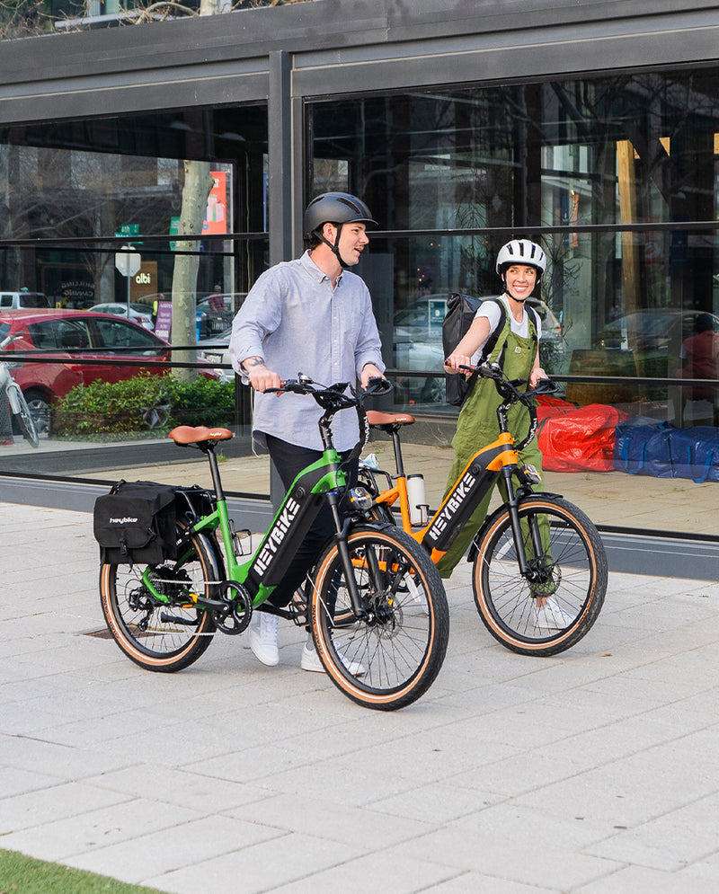 Two people, a man and a woman, wearing helmets and smiling while standing with HeyBike- Cityrun electric bicycles featuring lithium-ion batteries in an urban environment.