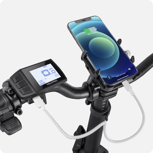 Close-up view of a Velotric - Fold 1 eBike handlebar fitted with a smartphone holder, a digital display screen, and a charging cable connected to the phone.