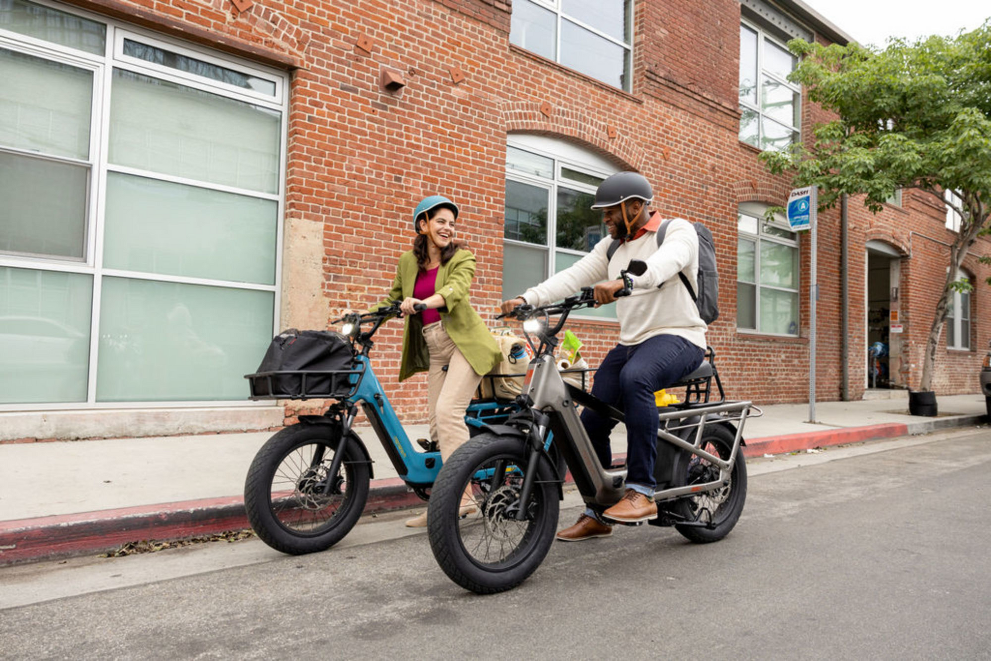 Two people riding Momentum - Cito E+ eBikes on a city street, smiling at each other, with brick buildings in the background.