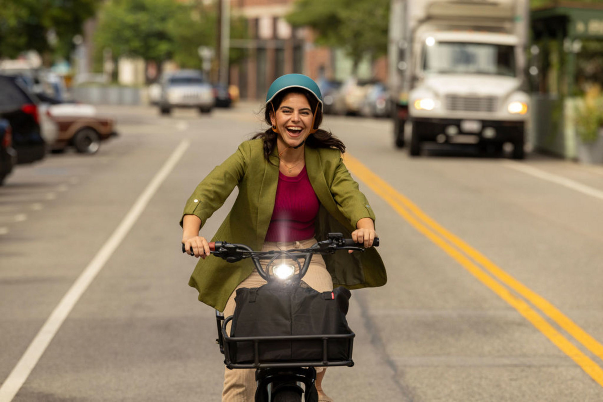 A joyful woman wearing a helmet rides a Momentum - Cito E+ electric bike down a city street, laughing with cars and a truck in the background.