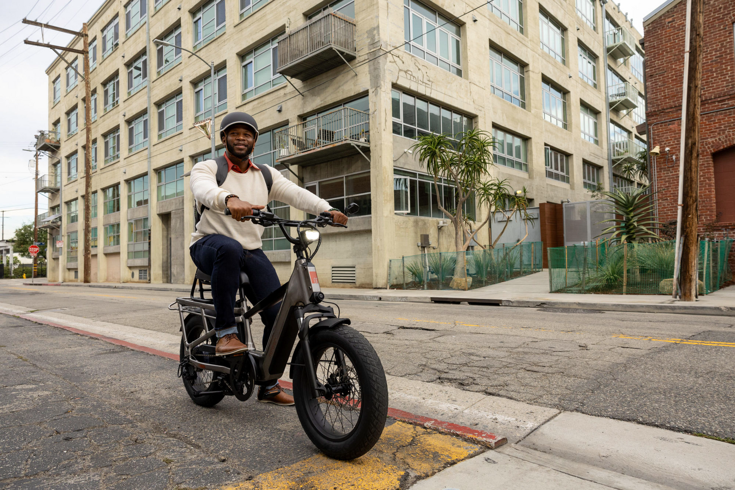 A man in a helmet riding a Momentum Cito E+ on an urban street with a large building in the background.