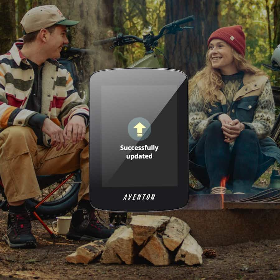 Two people sit by a campfire in the woods with their Aventon - Aventure 3 eBikes nearby. A digital screen in the foreground displays "Successfully updated.