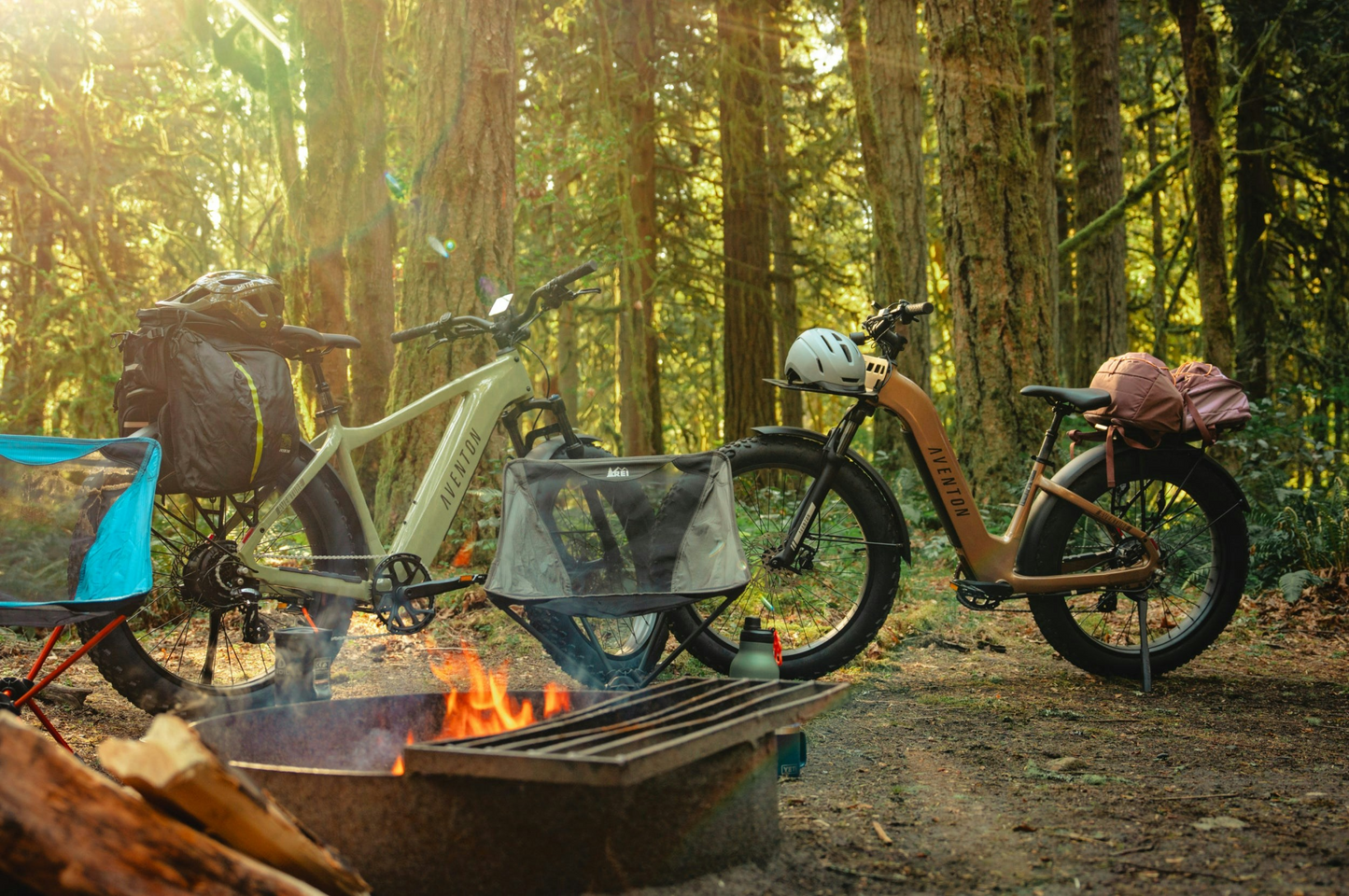 Two loaded bicycles, one being the Aventon - Aventure 3 from Aventon, are parked at a forest campsite near a campfire, gear and helmets attached, surrounded by tall trees with sunlight filtering through.