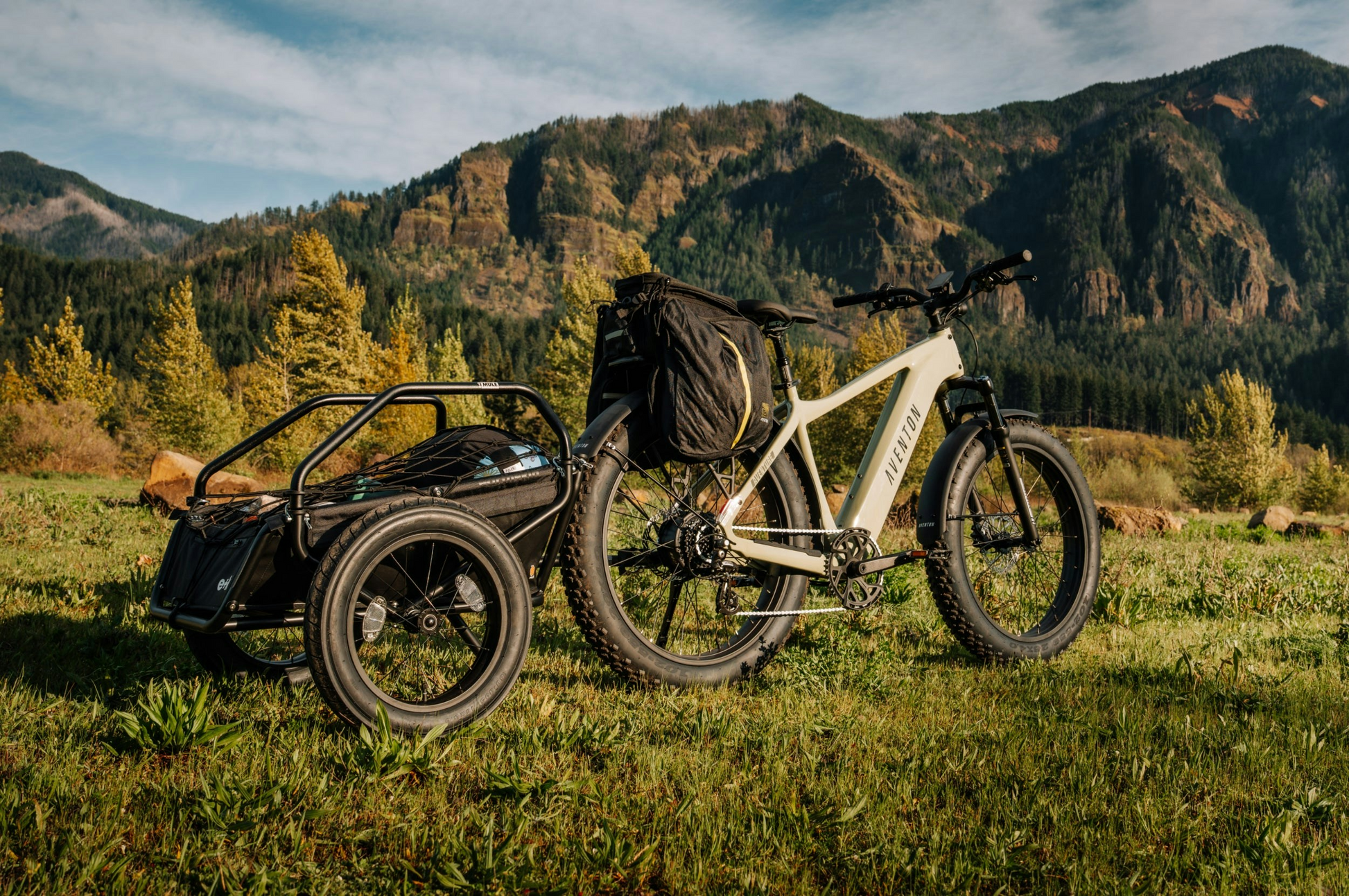 An Aventon Aventure 3 fat tire eBike by Aventon, equipped with a rear cargo trailer, is parked on grass with forested mountains in the background.
