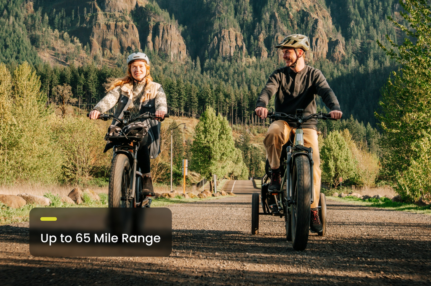Two people ride Aventon - Aventure 3 eBikes on a gravel path with trees and mountains, and a text overlay stating "Up to 65 Mile Range." Experience adventure and power with Aventon.