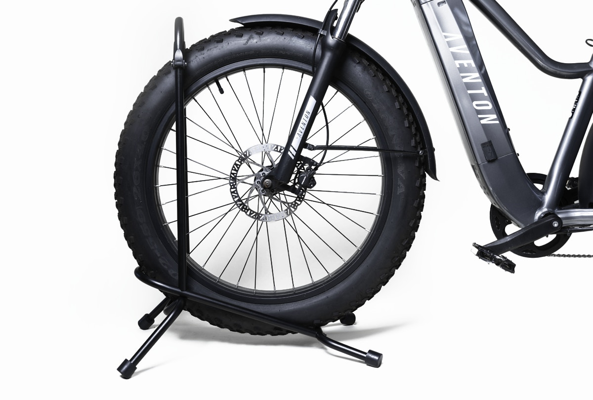 A front view of the black aluminum Aventon bike on an Aventon Floor Rack features wide tires, wheel, disc brakes, and part of the frame. The addition of a handy front basket enhances both functionality and style.